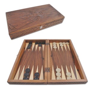 May include: A wooden backgammon set with a dark brown finish. The game board is open, displaying black and white game pieces. The lid, with a carved floral design, is positioned above the open board. A classic board game for two players.