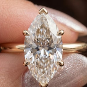 May include: A marquise-cut clear gemstone set in a polished yellow gold ring. The stone is secured by six prongs, with two at the top and bottom points and two on each side, featuring a faceted surface that reflects light in a brilliant pattern.