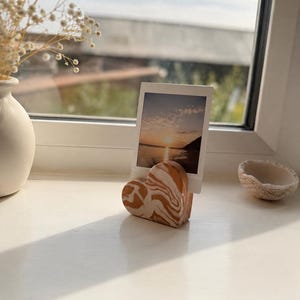 May include: A heart-shaped photo holder with a marbled pattern of burnt orange and white, featuring a vertical slot that holds a small rectangular photograph of a sunset over the ocean with a white border.