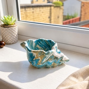 May include: A ceramic bowl shaped like a seashell with a marbled finish in shades of teal, white, and gold. The bowl has an irregular, wavy rim and a smooth, glossy surface. It is positioned on a white surface next to a small succulent plant.