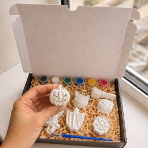 May include: A craft kit in a black box containing several white unpainted plaster figures including a pumpkin, cat, ghost, spider web, and broomstick, along with a set of six small paint pots and a blue-handled paintbrush for painting.
