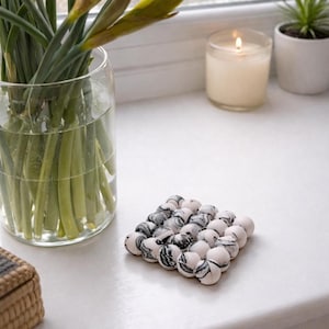 May include: A square coaster made of small, round, marble-patterned beads. The coaster is white with black and gray swirls. A glass vase with daffodils, a lit candle, and a small potted plant are in the background.