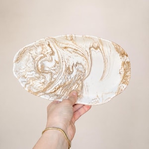 May include: An oval decorative plate with a marbled pattern in white and gold tones. The plate has an irregular shape with gently undulating edges. The plate is held up by a person's hand.