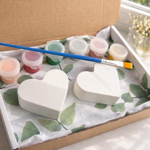 Könnte beinhalten: Ein DIY-Bastelset in einem Karton, mit zwei weißen herzförmigen Objekten, kleinen Farbtöpfen in verschiedenen Farben und einem blauen Pinsel. Das Set ist auf Papier mit Blättermuster angeordnet.