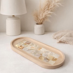 Peut inclure: Un plateau ovale en bois avec une incrustation de mosaïque. La mosaïque est composée de petits carreaux carrés irisés dans des tons crème et or. Le plateau est de couleur claire et repose sur une surface blanche. Une lampe et un vase blancs sont en arrière-plan.