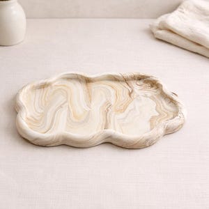 May include: A decorative cloud-shaped tray with a marbled pattern in shades of beige and brown. The tray has a wavy edge and is made of a solid material. The tray is designed for serving or display.