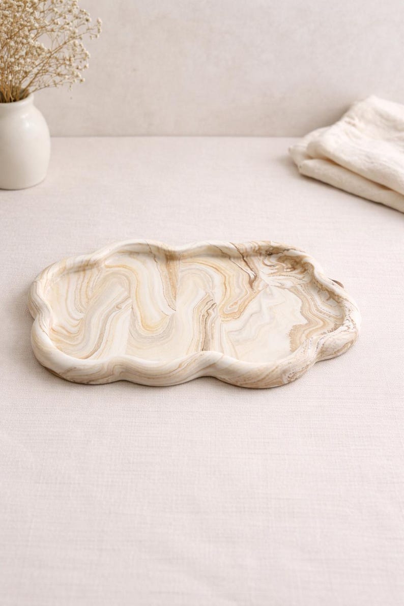 May include: A decorative cloud-shaped tray with a marbled pattern in shades of beige and brown. The tray has a wavy edge and is made of a solid material. The tray is designed for serving or display.