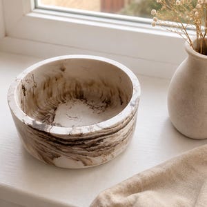 May include: A round decorative bowl with a marbled pattern in shades of brown and cream. The bowl sits on a white windowsill next to a small, beige vase holding dried flowers. The bowl's interior is a lighter cream colour.