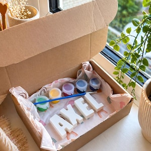Peut inclure: Un kit de bricolage dans une boîte en carton, avec le nom "AXEL" en lettres majuscules blanches. Le kit comprend de petits pots de peinture de différentes couleurs et un pinceau, le tout disposé sur du papier de soie à thème arc-en-ciel.