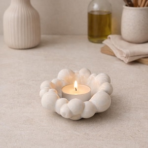 Op de afbeelding: Een witte, wolkvormige kandelaar met een brandende waxinelichtkaars. De houder heeft een textuur, bubbelachtig ontwerp. De achtergrond bevat een vaas met gedroogde bloemen, een fles olie en keukengerei.