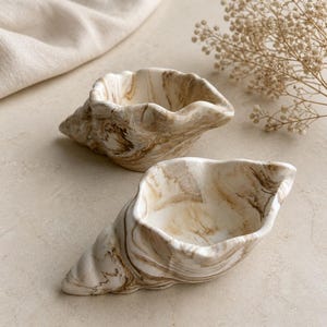 May include: Two shell-shaped bowls featuring a marbled pattern in shades of cream, beige, and brown. The bowls have a smooth, curved interior and a textured, spiraled exterior, mimicking the natural form of a seashell with a glossy finish.