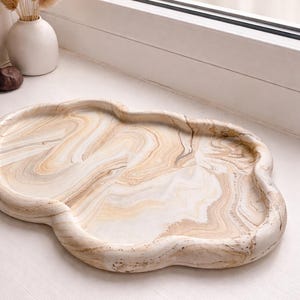 May include: A decorative, cloud-shaped tray with a marbled design in shades of beige and white. The tray has a smooth, glossy finish and a unique, organic shape. Perfect for displaying small items or serving snacks.