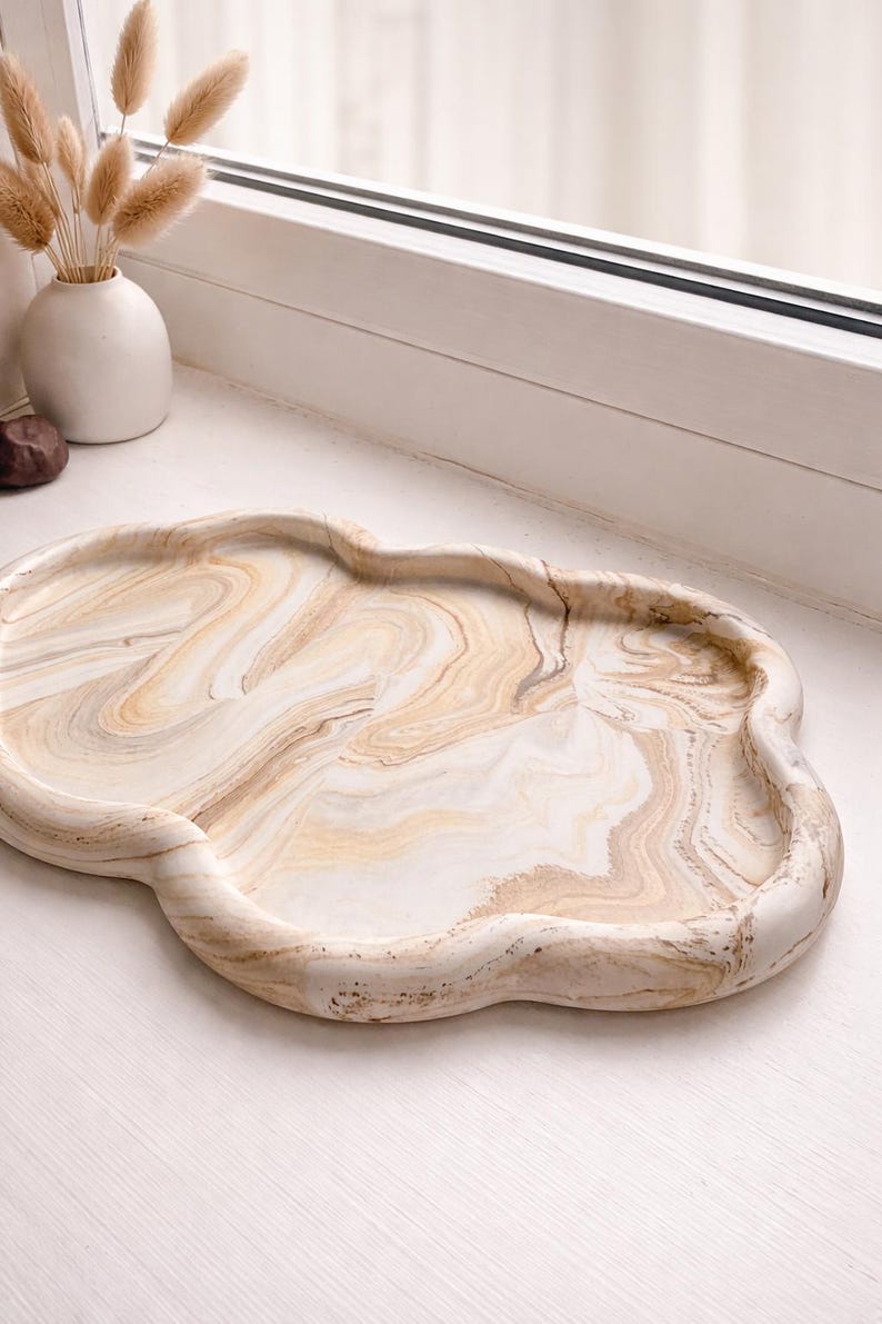 May include: A decorative, cloud-shaped tray with a marbled design in shades of beige and white. The tray has a smooth, glossy finish and a unique, organic shape. Perfect for displaying small items or serving snacks.