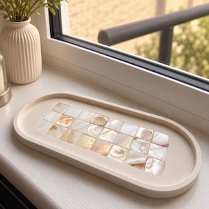 Puede incluir: Una bandeja ovalada de color blanco roto con una incrustación rectangular central compuesta por pequeñas baldosas cuadradas de nácar iridiscente. Las piezas muestran tonos variados de crema, beige y rosa suave con un acabado natural brillante.