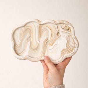 Puede incluir: Una bandeja decorativa con forma de nube con un diseño veteado en tonos blancos y beige. La bandeja está sostenida por una mano, mostrando su forma única y variaciones de color. La bandeja está hecha de un material sólido.