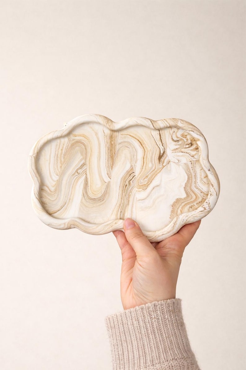 May include: A cloud-shaped decorative tray with a marbled design in shades of white and beige. The tray is held by a person's hand, showcasing its unique shape and color variations. The tray is made of a solid material.