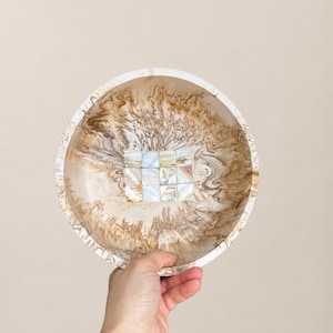 May include: A decorative bowl with a marbled pattern in shades of brown and cream. The bowl has a square mosaic tile design in the centre. The rim of the bowl is white. The bowl is held by a hand with a gold bracelet.