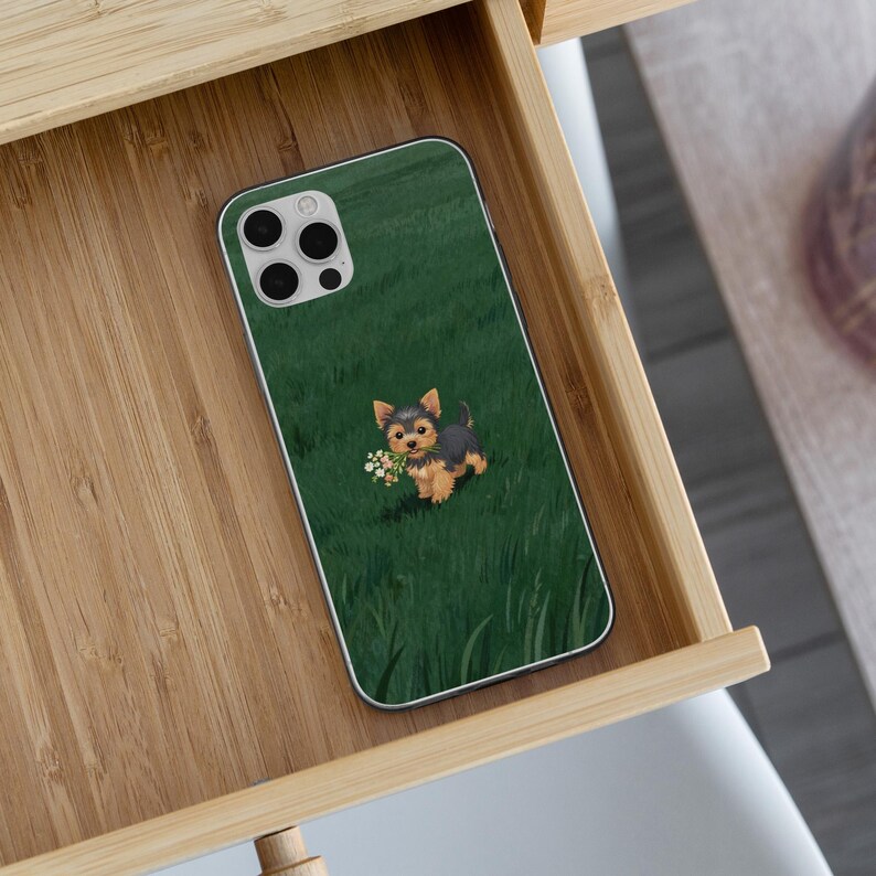 May include: A smartphone case featuring a Yorkshire Terrier puppy holding a bouquet of white flowers, set against a green grassy background. The case has a silver border and is resting in a wooden drawer.