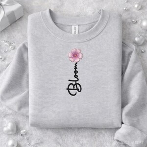 Peut inclure: Un sweat-shirt gris clair avec une fleur rose et le mot "Bloom" en écriture noire. Le sweat-shirt est plié, avec le motif centré sur le devant. L'étiquette de la marque Gildan est visible au dos du col.
