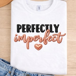May include: A white t-shirt featuring the phrase "PERFECTLY imperfect" in black and rose gold lettering, with a heart graphic. The shirt is folded on a light wooden surface, with denim jeans visible.