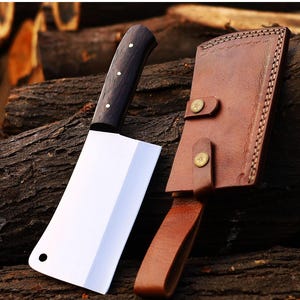 May include: A large cleaver knife with a dark wooden handle and a bright, polished blade. The knife is accompanied by a brown leather sheath with decorative stitching and brass-colored button closures. The knife and sheath are displayed on a background of stacked wood.