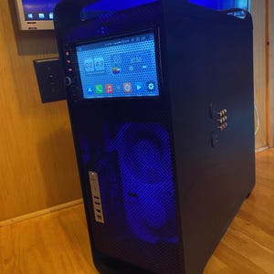 May include: A black computer tower with a built-in screen displaying a clock and app icons. The front panel has a mesh design with blue internal lighting. It features a handle on top and multiple USB ports.