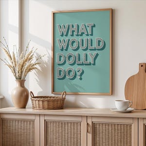 May include: A framed wall art print with a teal background featuring the white text WHAT WOULD DOLLY DO? in a marquee style font with small dots. The letters have a dark grey drop shadow, creating a three-dimensional effect on the poster.