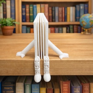 Può includere: Un oggetto bianco a forma di libro stampato in 3D, con braccia e gambe, seduto su una superficie di legno. L'oggetto ha una serie di pagine verticali ed è posizionato davanti a una libreria piena di libri colorati. Le braccia e le gambe sono articolate.