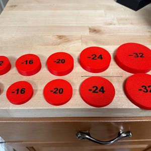 May include: Several red, circular objects with black numbers, ranging from -12 to -32, printed on them. They are arranged on a light-coloured wooden surface. Measurements are in centimetres.