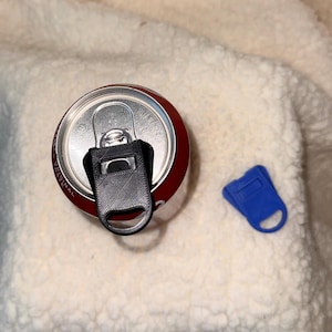 May include: A top-down view of a red and silver soda can with a black can opener attached to the tab. A separate blue can opener is next to the can. The can and openers are on a white, textured surface.