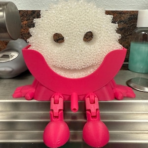 May include: A pink 3D-printed sponge holder shaped like a smiling character. The holder has a curved top, two eye-shaped holes, and two legs with feet. A white sponge is placed in the holder.