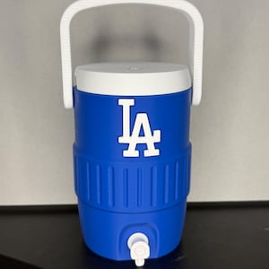May include: Blue and white water cooler with a white handle. The cooler has the letters "LA" in white on the front. The cooler has a white lid and a white spigot at the bottom. The cooler is sitting on a black surface.
