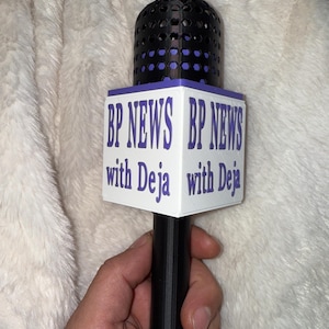 May include: A black and purple microphone with the text "BP NEWS with Deja" printed on a white rectangular base. The microphone is held in a hand against a fluffy white background.