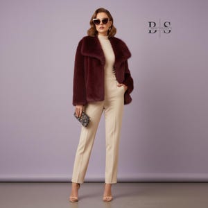 May include: A burgundy faux fur jacket with a wide collar is paired with cream-colored pants and a matching turtleneck. The outfit is accessorized with a patterned clutch and sunglasses. The Bella Solstice logo is visible in the upper right corner.