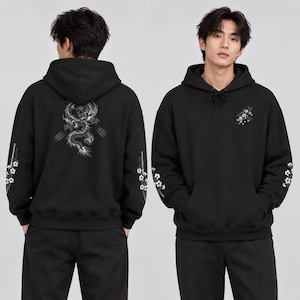 May include: A black hooded sweatshirt featuring a white dragon graphic on the back, a small floral design on the front chest, and white floral and sword motifs printed along both sleeves. The hoodie includes a drawstring at the neckline.