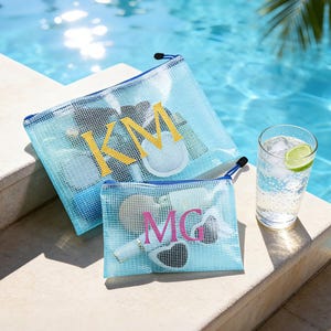 May include: Two blue mesh zipper pouches with personalised initials in yellow and pink. The pouches are filled with various items and are on a stone surface beside a pool. A glass of water with ice and a lime wedge is also present.