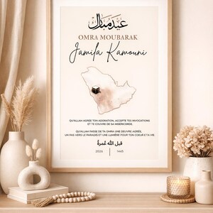 Peut inclure: Impression d'art encadrée avec calligraphie arabe et texte anglais. L'impression présente les mots "Omra Moubarak" et "Jamila Kamouni", ainsi qu'une carte de l'Arabie Saoudite. L'œuvre est exposée sur une étagère en bois avec des vases décoratifs.