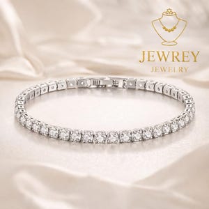 Op de afbeelding: Een zilveren tennisarmband met een rij ronde, heldere edelstenen. De armband is gemaakt van vierkante schakels en heeft een sluiting. De afbeelding bevat ook de tekst "JEWREY JEWELRY" en een logo.