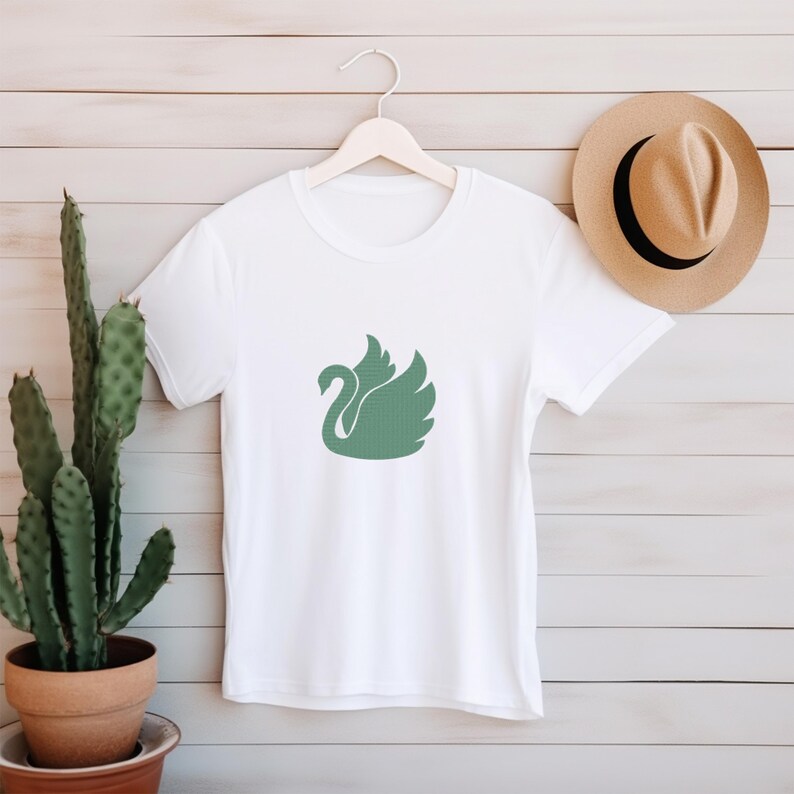 May include: White t-shirt featuring a green swan emblem. The swan design is centrally placed. A tan hat and a cactus in a brown pot are also visible. The t-shirt is hung on a wooden hanger.