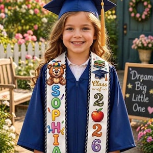 May include: A young graduate in a navy blue cap and gown, smiling at the camera. The graduation sash features a teddy bear, the name "Sophia", and the year "2026". The background includes a white picket fence and colorful flowers.