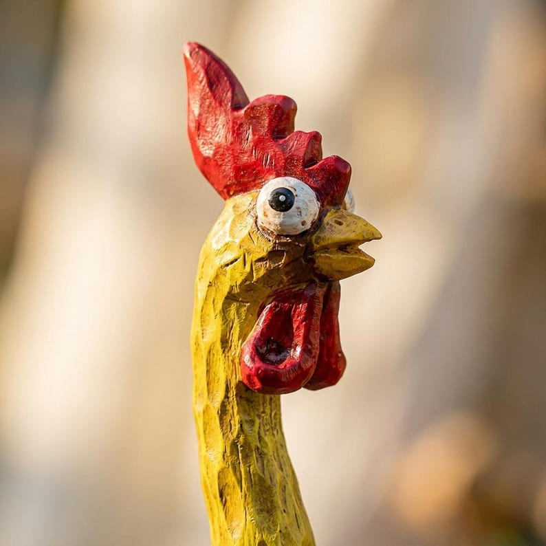 May include: A hand-carved wooden chicken figurine featuring a bright yellow neck and head, a vibrant red comb and wattle, and a yellow beak. The figure has a wide-eyed expression with large white eyes and black pupils, showing visible wood grain texture.