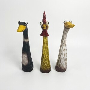May include: Three tall, slender carved figurines featuring a black duck with a yellow beak, a yellow rooster with a red comb, and a white duck with a yellow beak. Each figure has a textured, hand-carved surface and wide, expressive googly eyes.