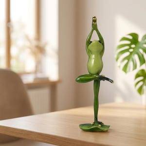 Puede incluir: Una figura de rana verde de pie sobre una base de nenúfar en una postura de equilibrio. La rana tiene los brazos levantados sobre la cabeza con las palmas juntas y una pierna doblada con el pie apoyado contra el muslo interior opuesto.