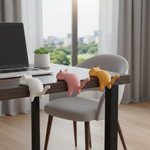 Puede incluir: Tres organizadores de escritorio con forma de gato en blanco, rosa y amarillo. Las figuras de gato están colocadas en el borde de un escritorio marrón oscuro, con sus cuerpos colgando por el costado. Una computadora portátil y una pequeña planta en maceta también están en el escritorio.