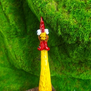 May include: A hand-carved wooden chicken figurine featuring a long, textured yellow body, a bright red comb and wattle, and two large, protruding white eyes with black pupils. The figure has a pointed beak and a smooth, tapered base.