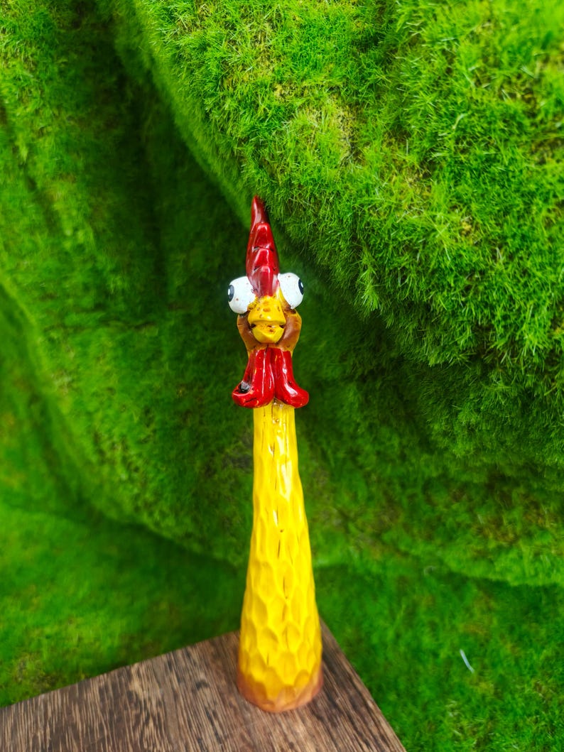 May include: A hand-carved wooden chicken figurine featuring a long, textured yellow body, a bright red comb and wattle, and two large, protruding white eyes with black pupils. The figure has a pointed beak and a smooth, tapered base.