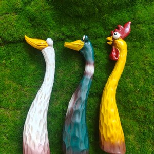 May include: Three hand-carved wooden bird figurines with elongated necks and textured surfaces. The left figure is white with a yellow beak, the center is teal with a yellow beak, and the right figure is yellow with a red comb and wattle.