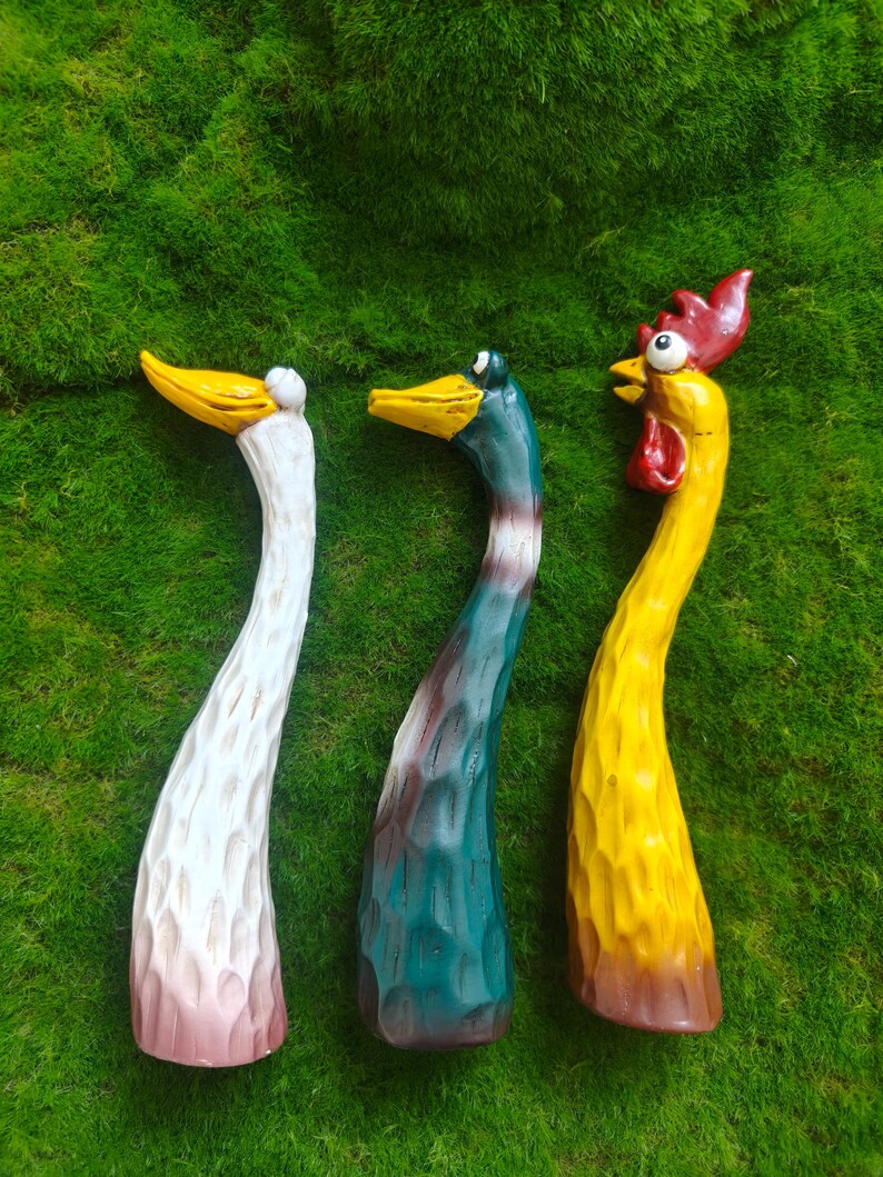 May include: Three hand-carved wooden bird figurines with elongated necks and textured surfaces. The left figure is white with a yellow beak, the center is teal with a yellow beak, and the right figure is yellow with a red comb and wattle.