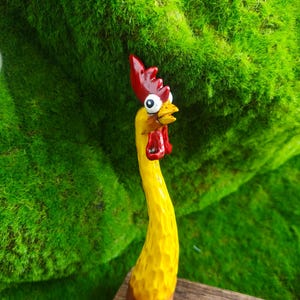 May include: A hand-carved wooden chicken figurine featuring a long, slender yellow neck and body with a textured surface. The chicken has a bright red comb and wattle, a yellow beak, and large, wide-eyed white eyes with black pupils looking upward.