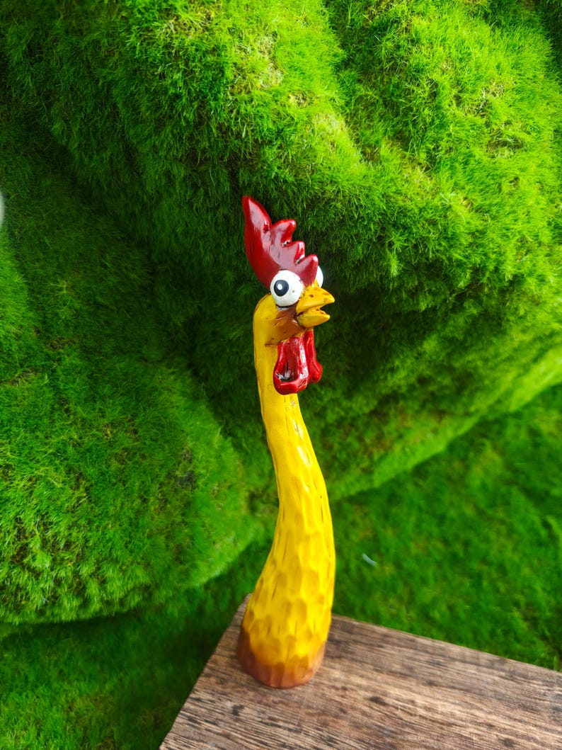 May include: A hand-carved wooden chicken figurine featuring a long, slender yellow neck and body with a textured surface. The chicken has a bright red comb and wattle, a yellow beak, and large, wide-eyed white eyes with black pupils looking upward.